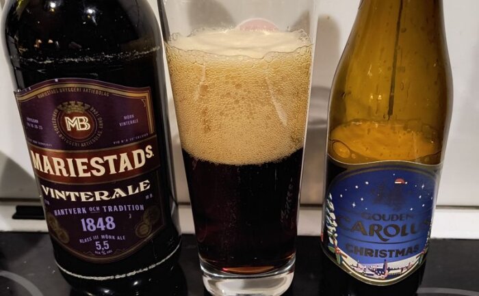 Flaskor av Mariestads och Gouden Carolus julöl med ett glas mörk öl mellan dem, upphällt med skum.