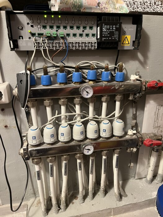 Fördelningssystem för golvvärme med termostater, rör och en cirkulationspump installerad på en vägg. Flera ventiler och en tryckmätare syns.