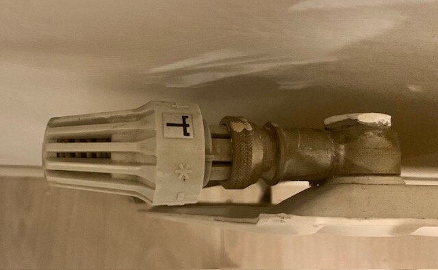 Termostat på element, visande en vit vred delvis mot en vägg, med en fastskruvad metallkoppling.