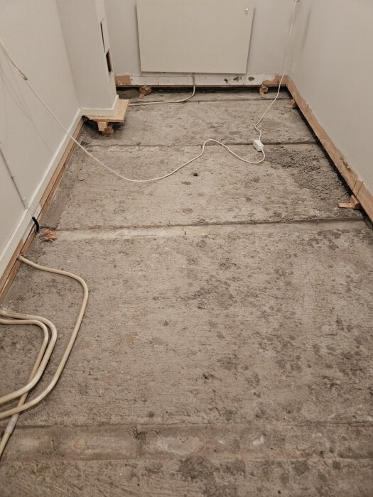 Utrivet betonggolv med synliga kanaler och borttagna spikreglar i ett rum där renovering pågår, med elsladdar längs väggarna.