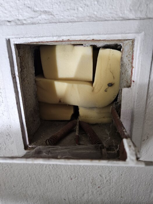 Öppet ventilationshål i en vägg, delvis blockerat med bitar av gul skumgummi och några synliga metallfjädrar, inramat av vitmålad metall.