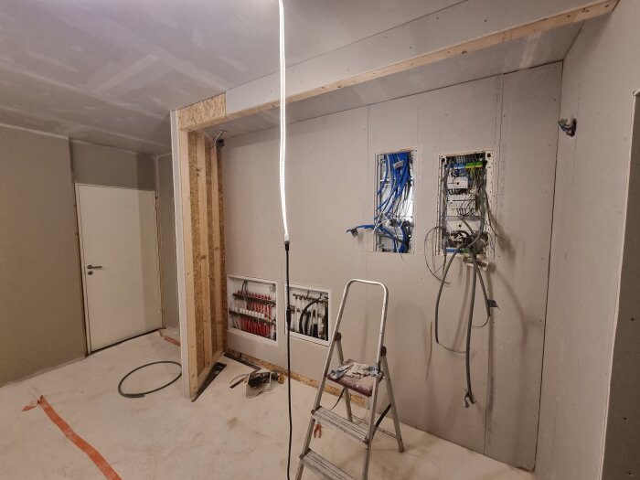 Renoveringsprojekt i hall med färdiggipsade och grundmålade väggar, synliga teknikluckor och verktyg samt stege på golvet.