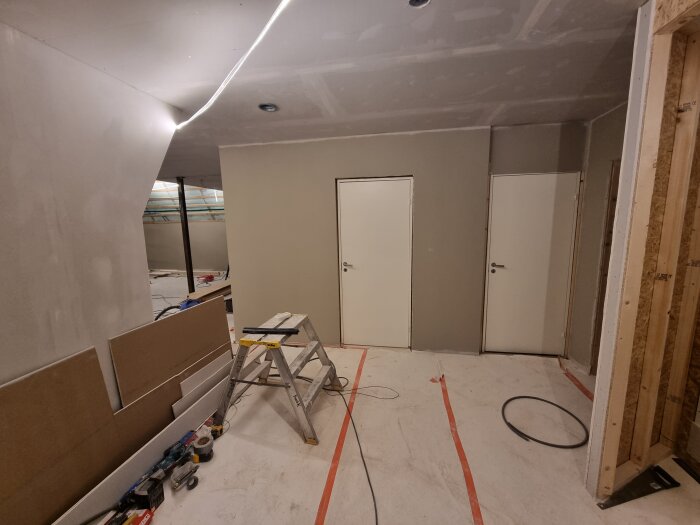 Rum under renovering med gipsade väggar, spackling, två vita dörrar och en stege. Byggmaterial ligger på golvet.