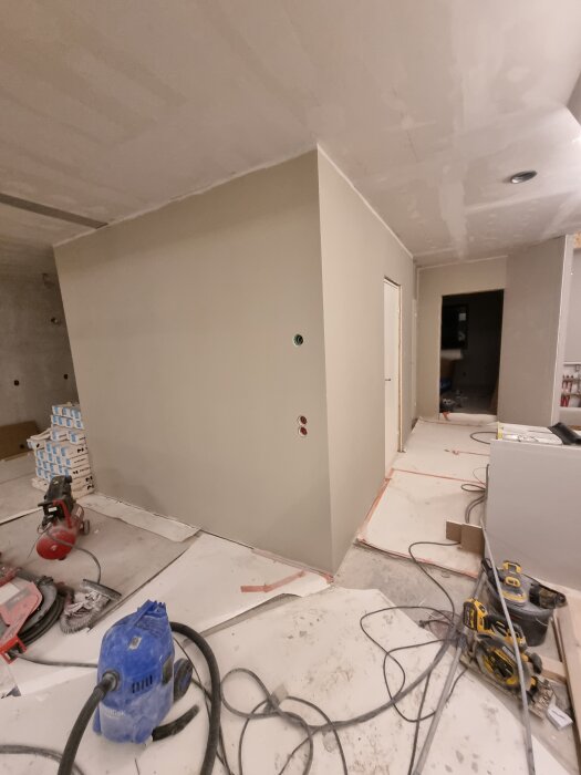 Beige gipsvägg i hall under konstruktion med verktyg och utrustning på golvet; eluttag synliga på väggen.