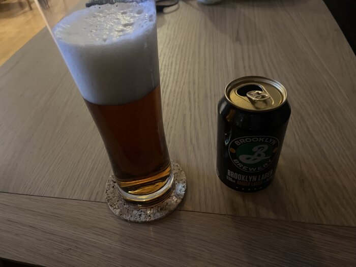 Ölglas med skum bredvid en öppnad burk Brooklyn Lager på ett träbord.