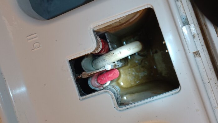 Öppen vit lucka med rött plugg och rör inuti en tank eller behållare, vatten synligt på botten. Pluggen verkar ha lossnat.