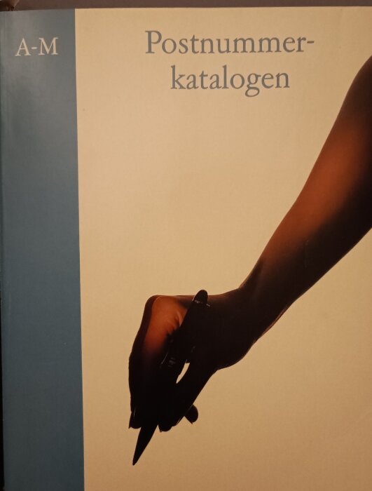 Framsidan av postnummerkatalogen A-M med en bild av en hand som håller i en penna.