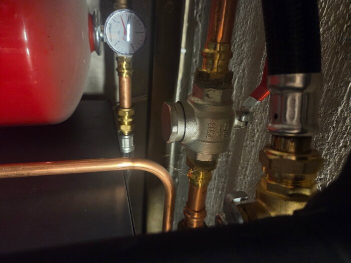 Tryckmätare och rör i ett värmesystem med kopparledningar, ventil och en röd tank synliga mot en vägg.