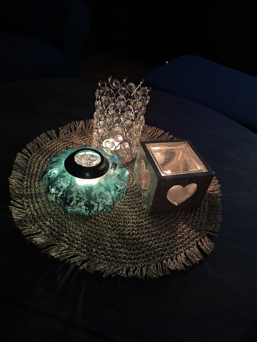 Tre ljusstakar med tända ljus står på en vävd rund bordstablett. Ljushållarna är av glas och metall, en med hjärtformad design.