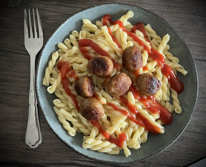 Pastatallrik med köttbullar och ketchup, serverad med en gaffel på träbord.