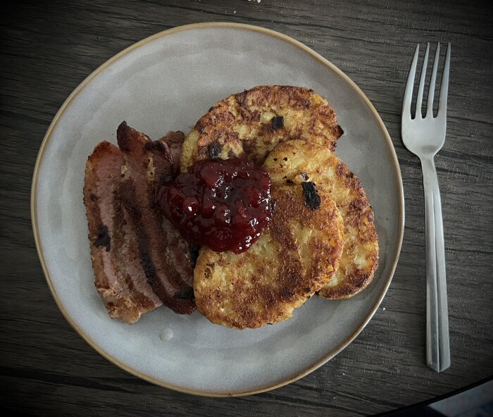 Potatisbullar med lingonsylt och stekt fläsk på en tallrik med en gaffel bredvid.