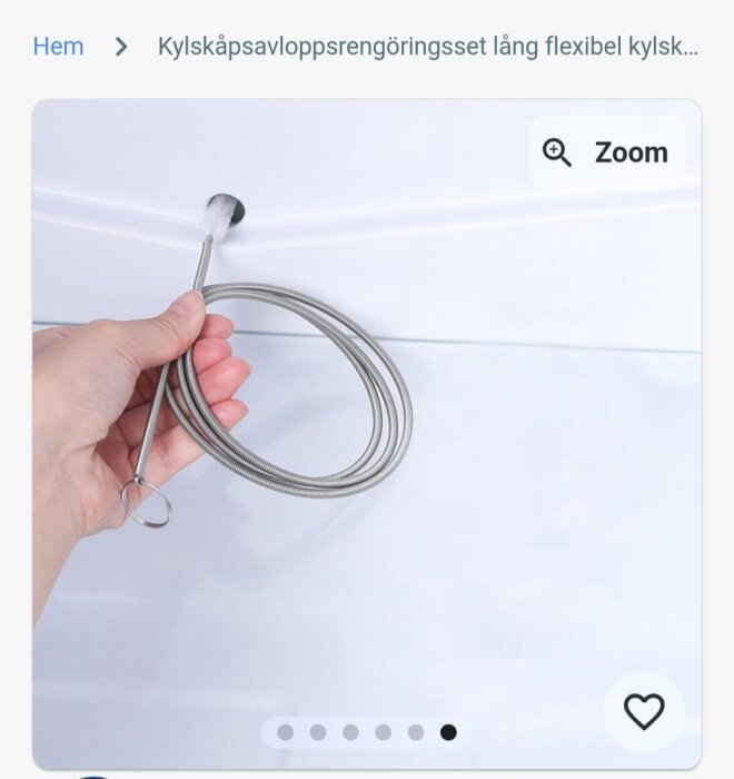 Hand håller en kylskåpsrengöringsståltråd med en borste i ena änden, används för att rensa stopp i kylskåpets kondensavlopp.