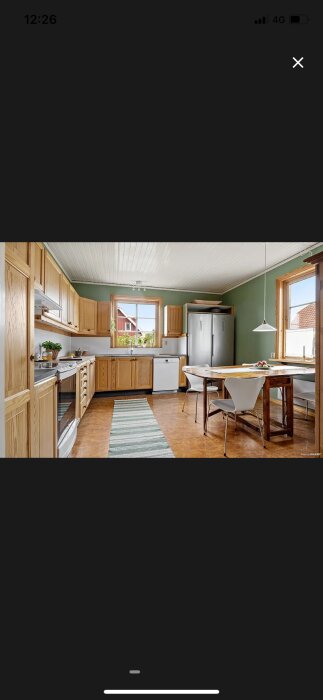 Kök med gröna väggar, trämöbler och två fönster. Matplats och kylskåp syns, samt dekorativa växter på fönsterbrädor.