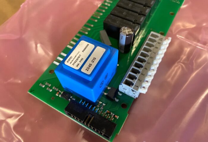 Helt nytt Nibe reläkort för bergvärmepumpar, med märkning och komponenter synliga på det gröna kretskortet, placerat på rosa underlag.