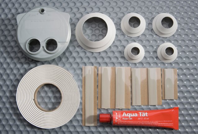Tätningskit för diskmaskinsgenomföringar med rörgenomföringar, tätningsmaterial och en tub Aqua Tät på en grå bakgrund.
