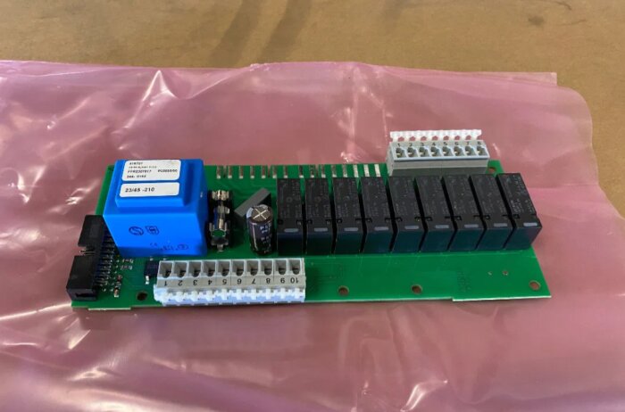 Nytt reläkort för Nibe värmepumpar, bergvärme. Grön kretskort med flera svarta reläer och en blå komponent, liggande på rosa underlag.