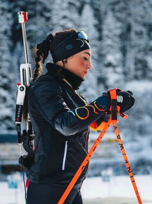 Skidskytt klädd i vinterkläder håller stavar och har en gevär på ryggen, snöig bakgrund.
