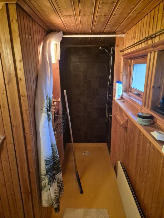 Badrum i fritidshus med träpanel, duschmodul med mörk vägg, plastmatta på golvet, duschdraperi och hylla med fönster vid sidan.