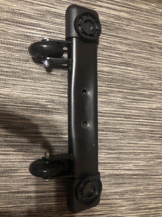 Svart metallfot till bänk med hjul, två hål i mitten och inklämd metall runt fästpunkterna.