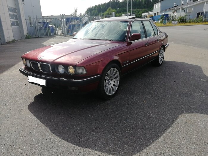 Röd BMW 750 från 1990 med V12-motor parkerad utomhus på asfalten, visas från sidan och en del av fronten.