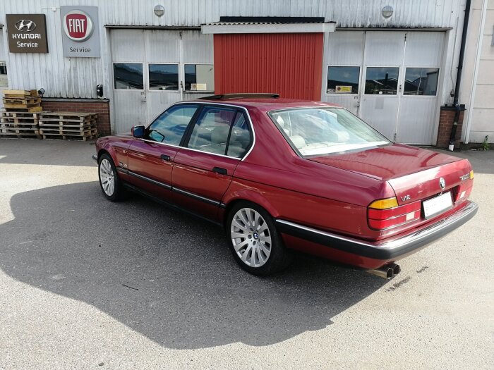 Röd BMW 750 från 1990 parkerad utanför en verkstad med Hyundai och Fiat-skyltar, sedd bakifrån med solens reflektion på karossen.