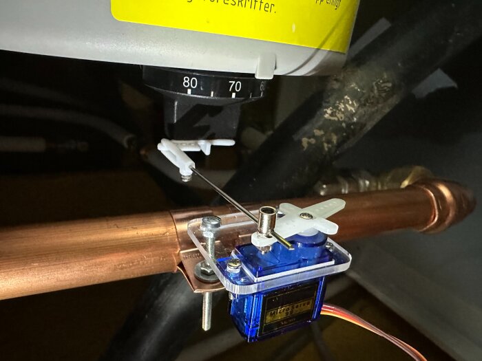 Termostatvred med servomotor och stötstång kopplad till en varmvattenberedare, monterad på en kopparrörsinstallation.