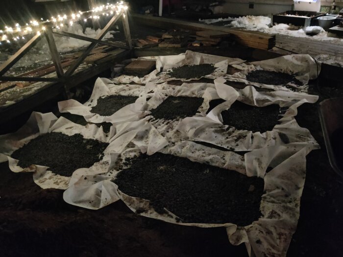 Makadam och markduk i grävda hål för altanbygge, omgiven av trädäck och ljusslinga i bakgrunden.