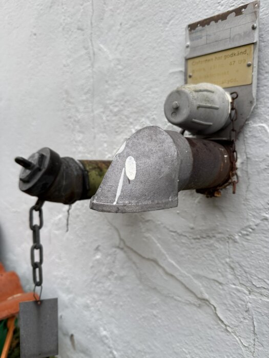 Svarta och grå rör på en vägg, det svarta med kedja och lock, det grå för avluftning bredvid en skylt med text.