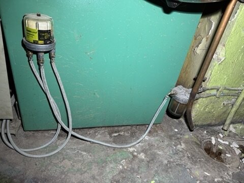 Elektrisk utrustning med anslutna kablar på en grön maskin, belägen nära ett kopparrör och ett hål i golvet.