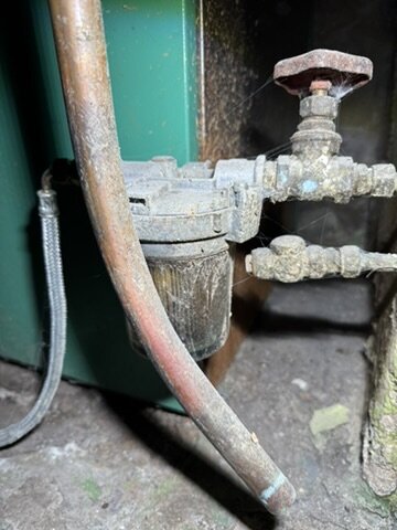 Rostiga rör och ventil ovanpå en gammal grön tank i en källare, används för vatten- eller värmesystemanslutning.