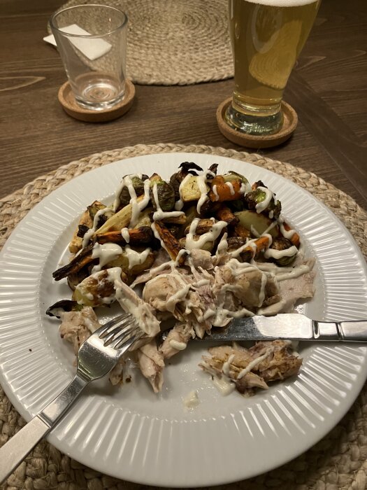 Tallrik med kycklingrester och rotfrukter täckta med vitlökssås; en gaffel och kniv ligger på sidan. Bakgrunden visar ett glas öl.