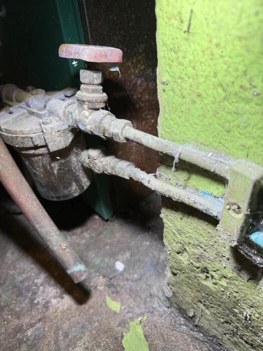 Närbild av en ledning med en ventil. Ledningen är fäst vid en grönmålad vägg och är dammig och sliten.