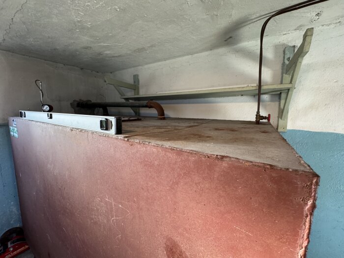 Röd tank med röranslutningar och en vattenpass på toppen, står i ett garage med blå och vit vägg.