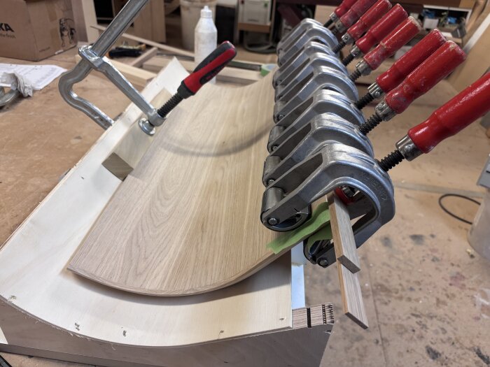 Pressar och limtvingar används för att böja och fästa en träfanér på ett böjt underlag i ett verkstadsprojekt.