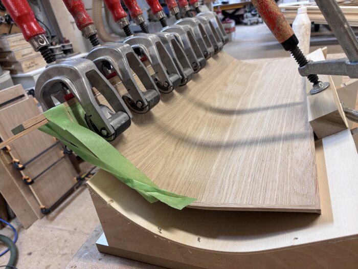 Flera tvingar håller fast trästycken under limning i ett snickeriprojekt. Gröna tejpbitar syns på toppen.