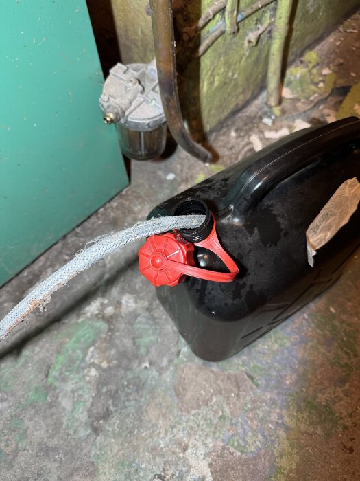 En dunk med svart vätska och en röd kork, ansluten till en pump genom en slang i ett rum med gröna väggar och rör.