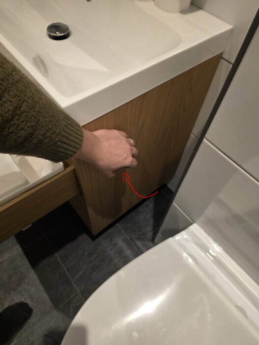Hand drar ut låda under handfatets skåp bredvid en toalettstol i ett badrum.