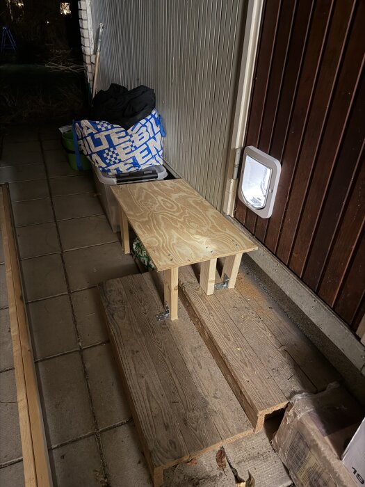Självbyggd trappa av träplankor leder till en kattlucka monterad i en trävägg, med en blå kasse och plastlåda intill.