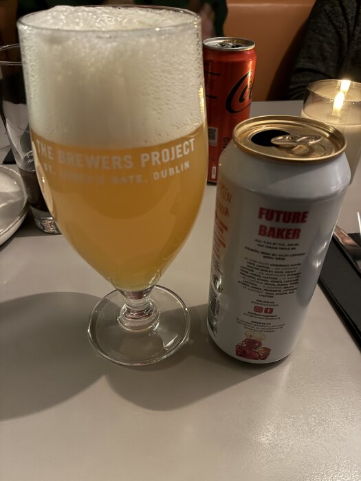 Ett glas öl från "The Brewers Project" och en burk med etiketten "Future Baker" står på ett bord bredvid ett tänt ljus och en läskburk.