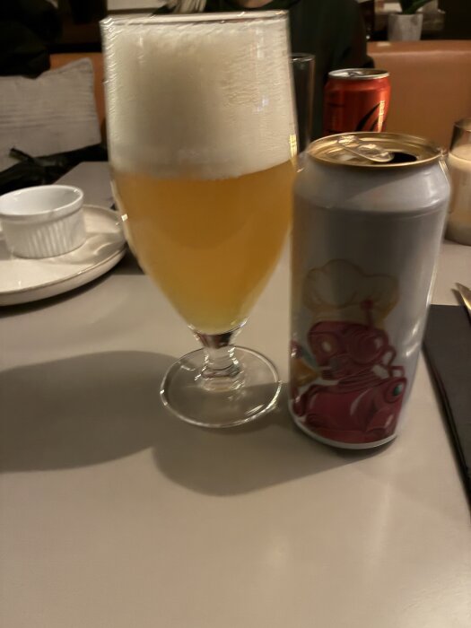 Ett glas öl med skum på toppen bredvid en ölburk med en tecknad figur, stående på ett bord i en restaurangmiljö.