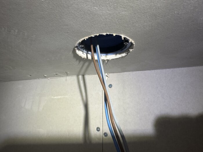 Öppning i taket med hängande elledningar i en dosa, utan synlig skyddsjord, mot en beige vägg.