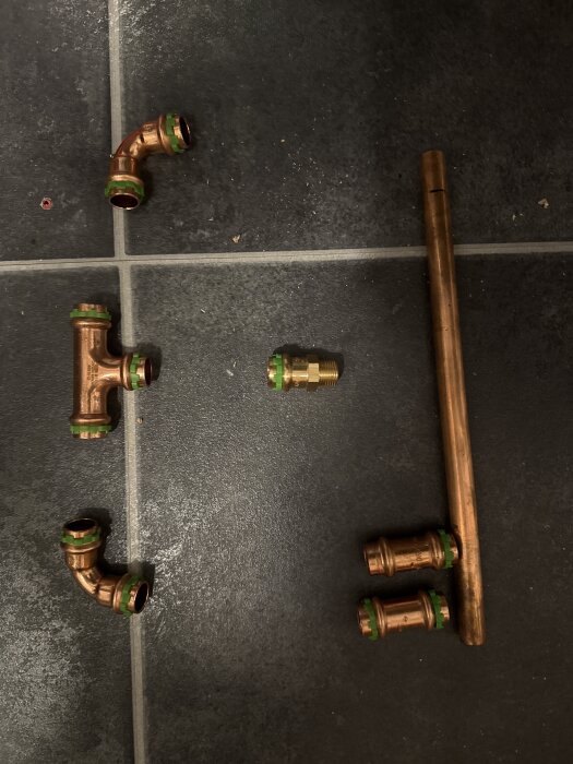 Kopparrör och kopplingar i olika former och storlekar ligger utspridda på ett mörkt klinkergolv.