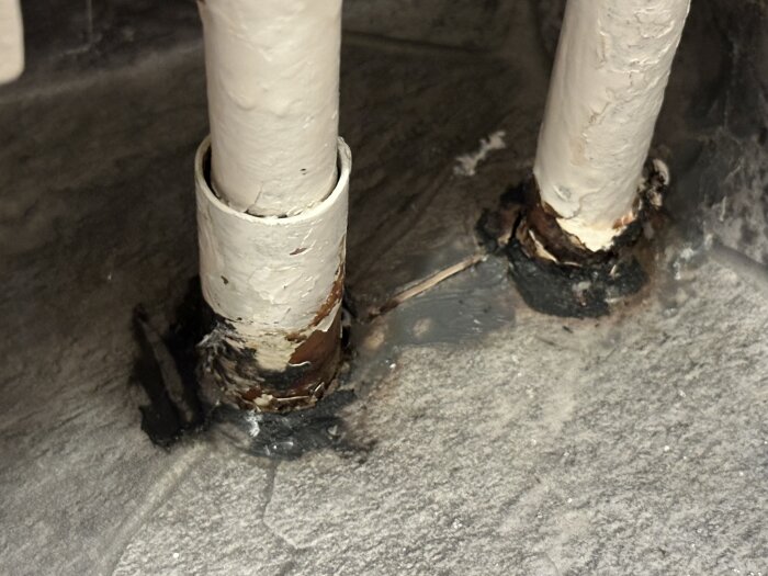 Närbild av två rörgenomföringar i ett badrumsgolv med svart massa kring basen och spricka i våtrumsmattan mellan rören.