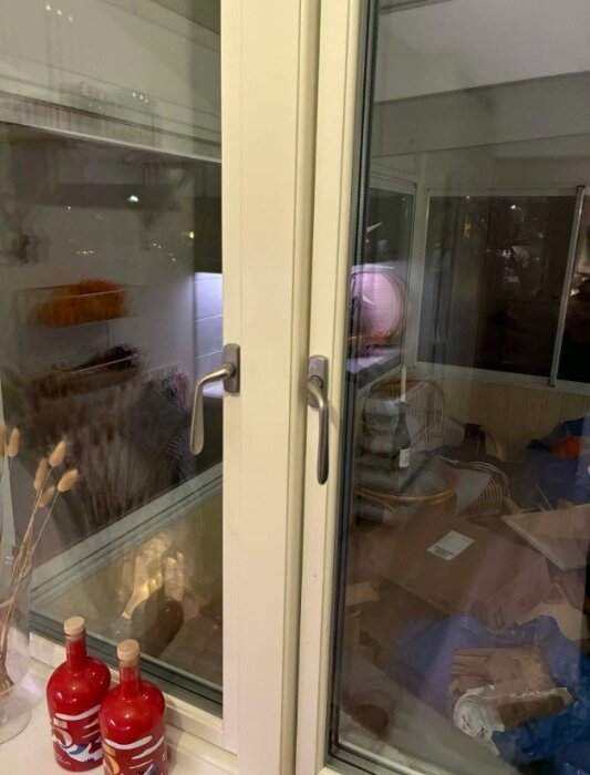 Moderna fönster med enkla metallhandtag och utsikt mot ett rum med inredningsdetaljer, inkl. två röda flaskor på fönsterbrädan.