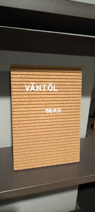 Skylt med texten "Väntöl 56 KR" i vitt på en brun korkbakgrund.