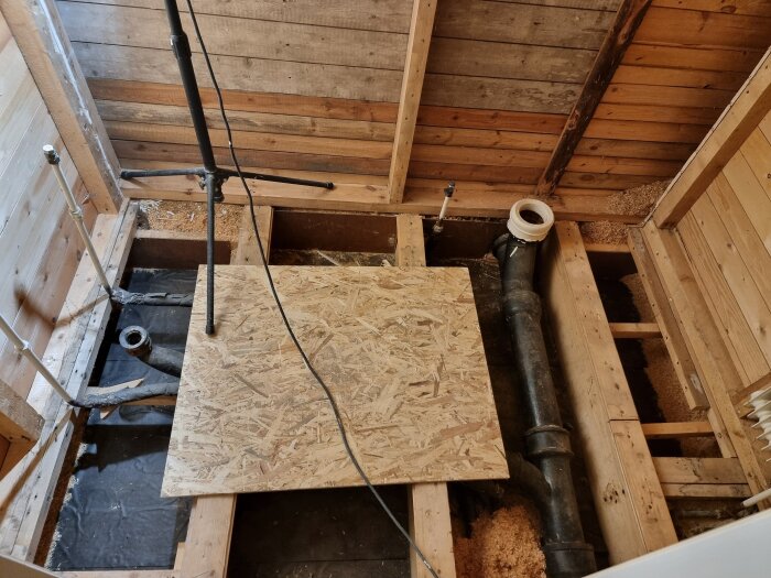 Renoverat badrumsgolv med exponerade rör och träbjälkar, samt en plywoodskiva och ett stativ i bild.