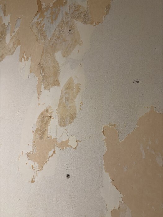 Vägg med delvis borttagen strukturtapet, fläckar av brunt papper och rester av tapet kvar. Väggen är ojämn och saknar primer på vissa ställen.