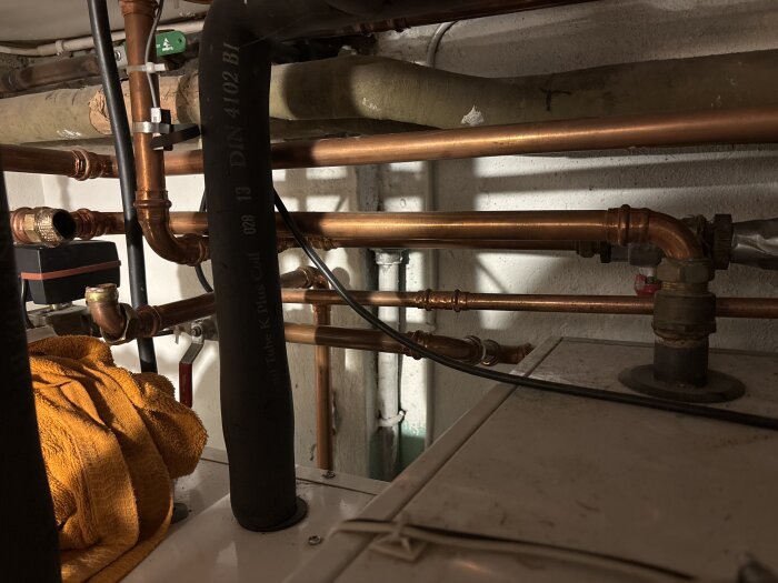 Kopparrör och VVS-installation i källarmiljö med isolerade rör och en orange trasa i förgrunden.