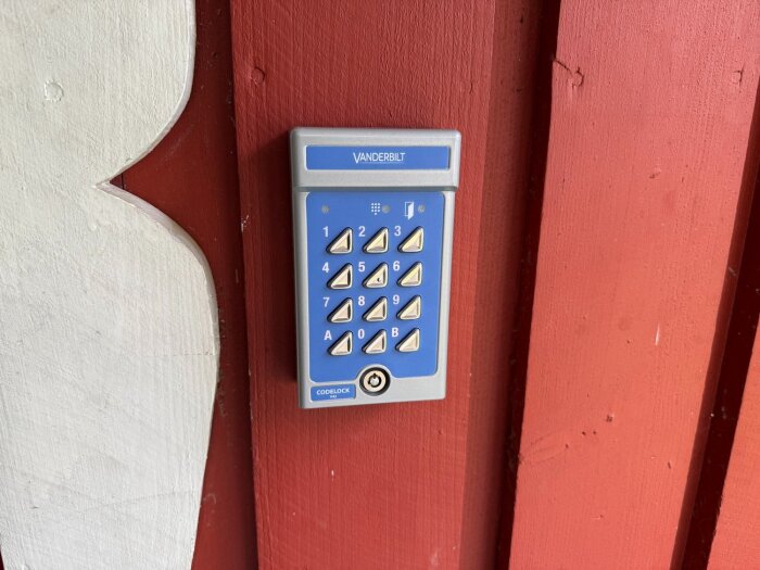 Grå-blå kodpanel från Vanderbilt monterad på ett rött träparti, används för säker åtkomst till husets bakdörr.