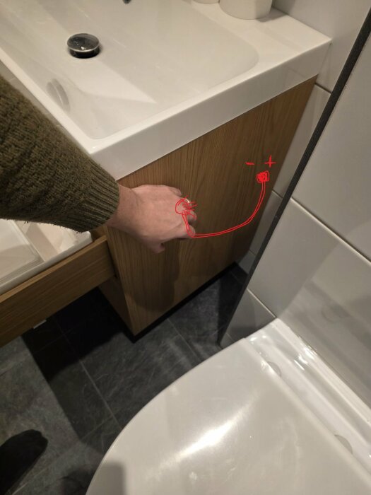 Hand pekar mot badrumskåp med handritade röda linjer som visar potentiell dragning för toalettduschens blandare.